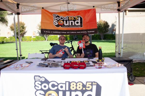 Byron the Curator and a SoCal Sound volunteer sitting at The SoCal Sound booth with various SoCal Sound merchandise.